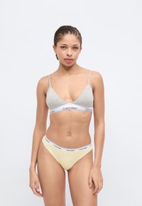 Γκρι βαμβακερό τρίγωνο σουτιέν με λεπτές τιράντες και λευκή ταινία, συνδυασμένο με κίτρινο βαμβακερό στρινγκ με λευκή ζώνη μέσης που φέρει την ετικέτα "Calvin Klein".