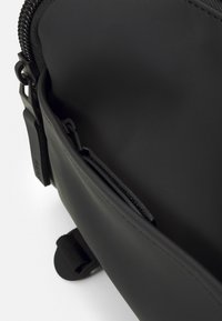 Borsa nera realizzata in materiale liscio, con una tasca con zip e accenti testurizzati. Il design include linee minimaliste e componenti robusti.