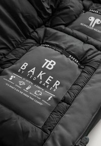 Zwarte pufferjack met een gequiltte textuur en een grijs label waarop de merknaam "Baker" en de verzorgingsinstructies staan. Tot de kenmerken behoren waterbestendigheid en gerecycled vulmateriaal.