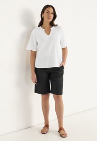 Vit, kortärmad, strukturerad topp med V-ringning; kombinerad med svarta knälånga shorts och bruna platta sandaler. Modell står med en hand i fickan.