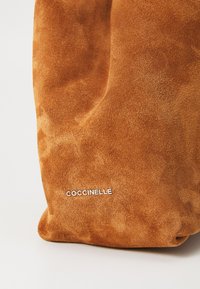 Braune Wildledertasche mit einer weichen Textur, ausgestattet mit einem auffälligen silbernen "Coccinelle"-Logo im unteren Vorderbereich.
