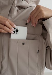 De handen van een persoon plaatsen een witte smartphone met drievoudige lenscamera in de borstzak van een beige jas met een drukknoop.