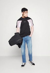 Camiseta preta com detalhes em blocos de cor rosa e branco, combinada com jeans azul claro e ténis pretos. Segurando um casaco preto.