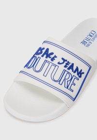 White slide sandal with a textured rubber sole. Features a wide strap adorned with blue "Versace Jeans Couture" lettering and border.