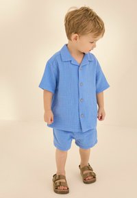Conjunto de algodón azul claro que incluye una camisa de botones de manga corta y pantalones cortos a juego. Combinado con sandalias marrones y un acabado texturizado.