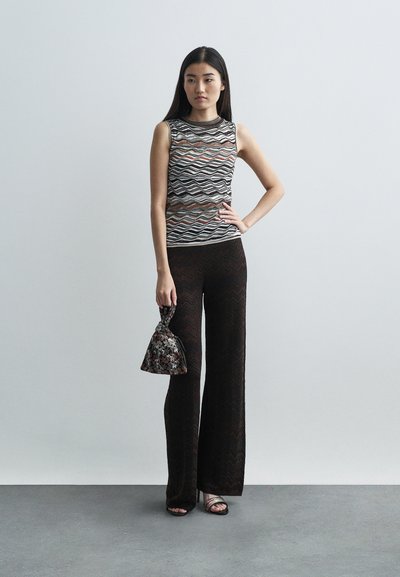 Γυναίκα που στέκεται με το ένα χέρι στη μέση, φορώντας ένα αμάνικο μπλουζάκι με σχέδια, σκούρους φαρδείς παντελόνες, σανδάλια και κρατώντας μια μικρή λουλουδάτη τσάντα.