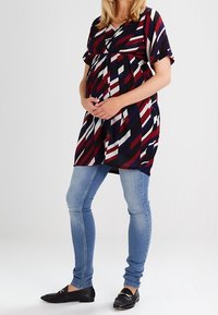 Raskausmekko tummansinisellä väriyhdistelmällä, jossa on punaisia ja valkoisia vinoraitoja, lyhyet hihat, ja kootut osat rinnan alta. Mekko on yhdistetty vaaleansinisiin farkkuihin ja mustiin loafereihin.