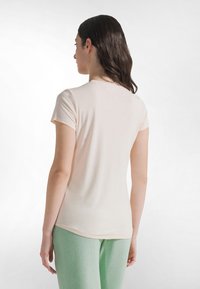 Jasnoróżowa koszulka z krótkim rękawem i okrągłym dekoltem, o gładkiej fakturze i lekko zaokrąglonym dole. Noszona z jasnooliwkowymi spodniami.