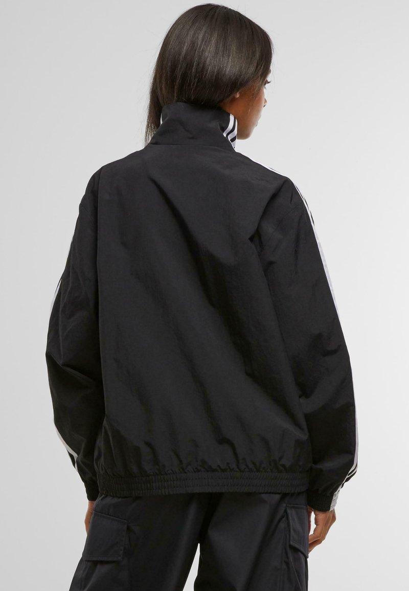Schwarze Nylonjacke mit hohem Kragen, elastischem Bund und weißen Seitenelementen, die einen lockeren Schnitt und eine glatte Textur aufweist.