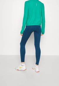 Maglietta verde a maniche lunghe con colletto alto, abbinata a leggings blu scuro. I leggings hanno una texture liscia e un design aderente. Sneakers bianche incluse.