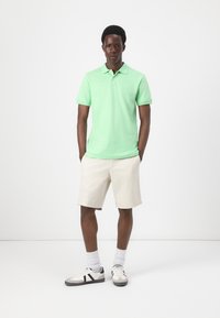 Green cotton polo shirt with a collar, beige shorts, and gray sneakers with black accents. Model stands straight with hands in pockets.