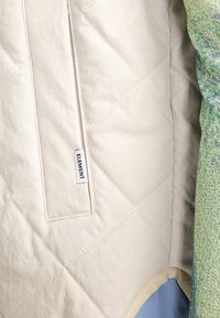 Intérieur d'une veste beige matelassée avec une poche à rabat, arborant une étiquette noire "ELEMENT". Doublure texturée colorée visible sur le bord droit.