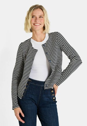 Cardigan rayé en bleu marine et crème, avec un col rond et une fermeture à boutons-pression. Porté par-dessus un débardeur blanc à côtes avec un pantalon en denim foncé.