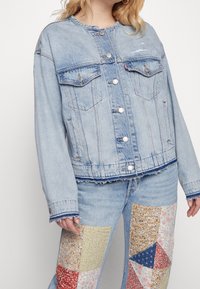 Světle modrá džínová bunda s límcem, předními kapsami a stříbrnými knoflíky. Obsahuje třepení na spodním lemu a je spárována s patchworkovými džíny v různých barvách.