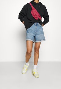 Sudadera negra, pantalones cortos de mezclilla azul claro, bolso de hombro rosa y zapatillas blancas/amarillas. Diseño simple con ajuste casual y pantalones cortos arremangados.