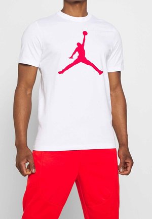 Mann trägt ein weißes T-Shirt mit großem rotem Jumpman-Logo und leuchtend roten Hosen, steht vor einem einfarbigen Hintergrund.