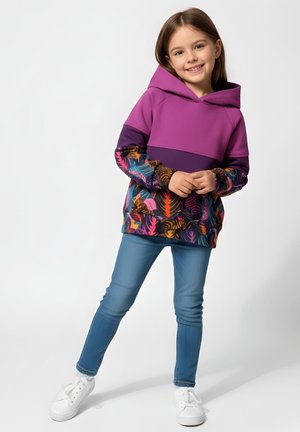 Uśmiechnięta dziewczyna w fioletowej bluzie z kapturem z kolorowymi wzorami liści, niebieskich dżinsach i białych tenisówkach stoi na jednolitym tle.