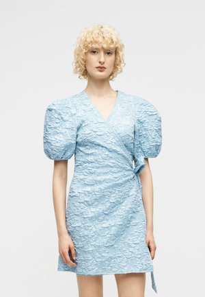 Kobieta w jasnoniebieskiej sukience kopertowej o fakturowanej tkaninie i obszernych bufianych rękawach, stojąca na tle jednolitego białego tła.