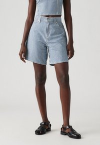 Blå och vita vertikala randiga högmidjade shorts med knappstängning och bältesöglor, tillverkade av denim. Svarta sandaletter med remmar kompletterar outfiten.