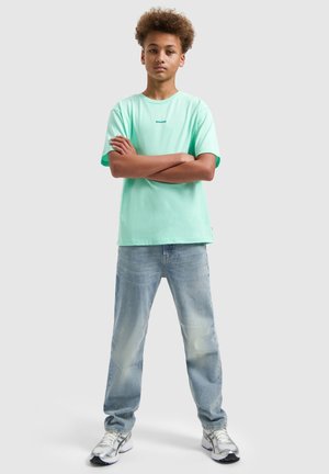 Teen boy with curly hair stands with arms crossed wearing a mint green t-shirt, light blue jeans, and white sneakers on a plain background.