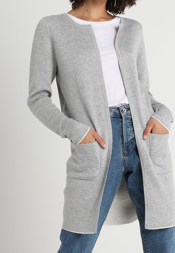 Femme portant un long cardigan gris clair ouvert à l'avant avec des poches, par-dessus un chemisier blanc et un jean bleu, les mains dans les poches.