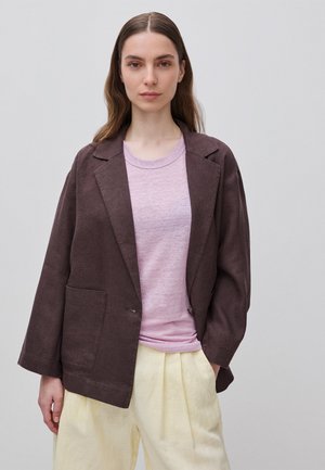 Femme aux longs cheveux bruns portant un blazer marron ample, un t-shirt rose clair et un pantalon large jaune pâle sur un fond gris uni.