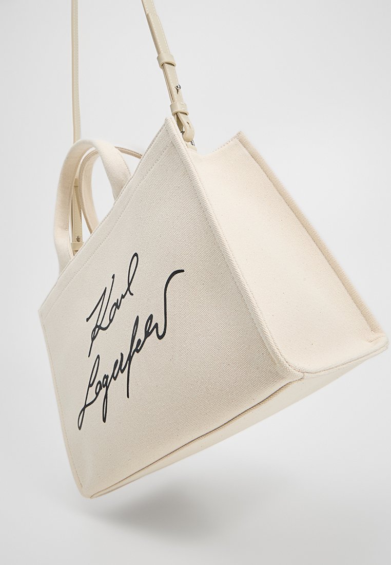 Sac fourre-tout en toile de couleur crème. Doté d'une forme structurée avec de courtes poignées et une longue bandoulière réglable. Orné d'une signature noire.