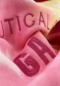 Tela rosa con texto bordado en un tono más oscuro de rosa. El material parece suave y tiene una ligera textura.