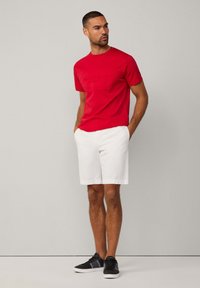Camiseta de algodón roja con el logo en relieve "HACKETT SPORT", combinada con pantalones cortos blancos y zapatillas negras. Corte casual con mangas cortas.