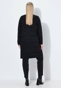 Dlouhý černý cardigan s texturovanou tkaninou, zdobený opakovaně embosovaným textem. Spárováno s černými kalhotami a kotníkovými botami. Jednoduchý design.