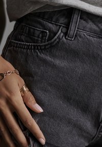 Jean en denim noir avec une finition texturée, dotés de poches avant et d'un ourlet effiloché. Une main repose sur le tissu, ornée de bagues en or.