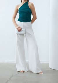 Teal top med en axel och rynkad textur, i kombination med högmidjade vita vida byxor. Håller i en silverglittrande klutch.