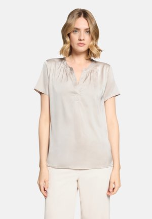 Woman with shoulder-length blonde hair wearing a short-sleeve, light beige satin blouse and white pants against a plain background.