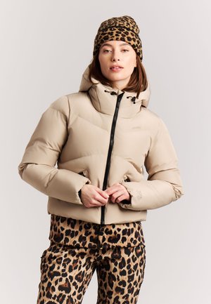 Vrouw die een beige donsjas met zwarte rits draagt, luipaardprintmuts en bijpassende luipaardprintbroek, staand tegen een effen achtergrond.