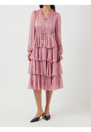 Kvinde iført en lyserød lagdelt midikjole med lange ærmer, flæset V-halsudskæring, knapper foran, taljebånd og sorte spidse hæle.