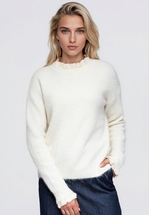 Une femme porte un doux pull blanc au col et aux poignets bordés de dentelle, associé à une jupe en jean foncé sur un fond uni.
