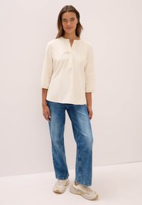 Donna in piedi con una blusa beige a maniche lunghe, jeans blu e sneakers beige su uno sfondo neutro semplice.