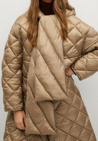 Poncho acolchado beige con capucha, que presenta patrones de costura diagonales y una textura suave y brillante. La tela parece ligera y cálida.