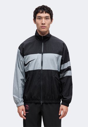 Junger Mann, der nach vorne schaut, trägt eine schwarz-graue Sportjacke mit Reißverschluss und Nike- sowie PSG-Logos auf schwarzen Hosen.