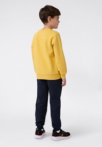 Geel sweatshirt met een ronde hals en ribgebreide boorden, gecombineerd met marineblauwe joggingbroek. Zwarte sneakers met oranje en groene accenten.