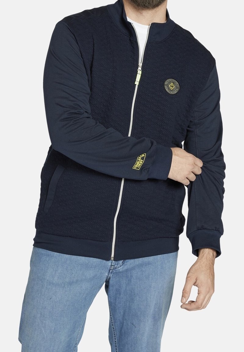 Homme portant une veste texturée bleu marine zippée avec des patchs sur la poitrine et la manche, associée à un jean bleu clair, ajustant sa manche gauche.