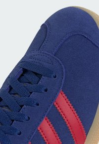 Nærbillede af en blå ruskinds sneaker med marineblå snørebånd, røde læderstriber og beige struktureret sål.