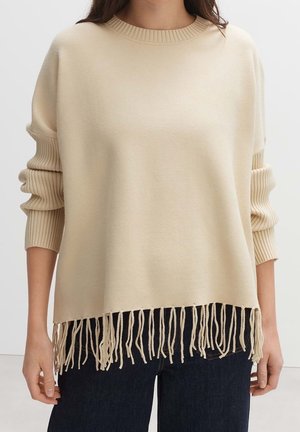 Woman wearing cream-colored knit sweater with ribbed sleeves and fringe hem paired with dark blue jeans.