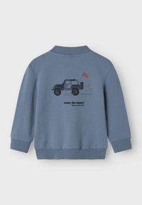 Zils džemperis ar ribotām piedurknēm un apakšmalu, uz kura attēlota džips un sarkana karoga. Teksts vēsta "ātrāk šķērsot tuksnesi rietumos".