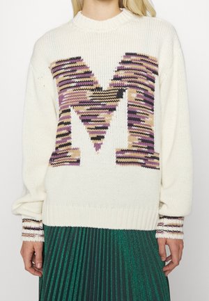 Femme portant un pull en maille crème avec une grande lettre rayée multicolore "M" et des poignets assortis, associée à une jupe plissée verte.