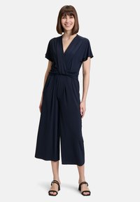 Tuta blu navy con scollatura avvolgente e design a gamba larga. Maniche corte, dettaglio in vita annodato, realizzata in tessuto morbido. Sandali neri con tacco.
