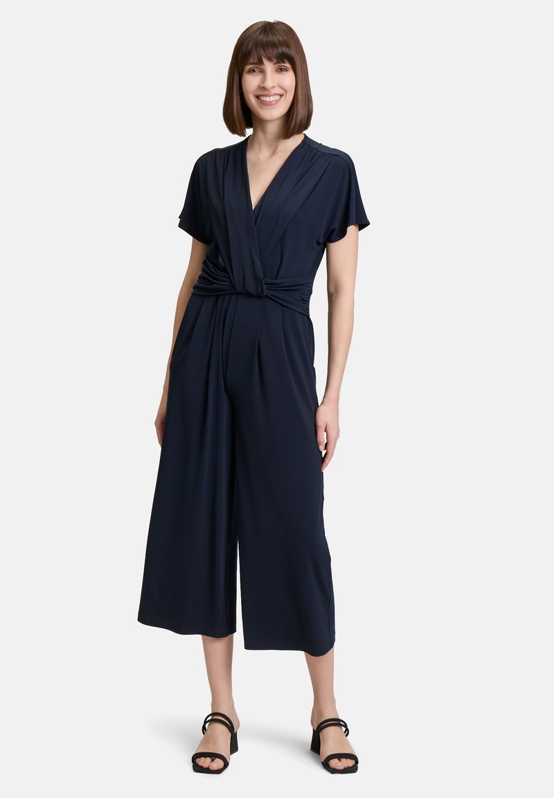 Tuta blu navy con scollatura avvolgente e design a gamba larga. Maniche corte, dettaglio in vita annodato, realizzata in tessuto morbido. Sandali neri con tacco.