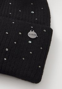 Bonnet en tricot noir avec une texture côtelée, orné de petits strass transparents. Comprend une étiquette ronde en argent gravée "GUESS."