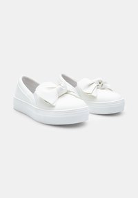 Fehér slip-on sportcipők, textúrált fehér gumitalppal, elöl egy nagy díszítő masnival és sima szintetikus felső résszel.