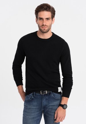 Suéter negro de punto con cuello redondo, mangas largas, de tela texturizada, combinado con jeans azules y un reloj negro, con una etiqueta en la cintura.